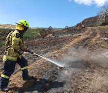 A firefighter in full PPE sprays the side of a hill, still smouldering from a fire.